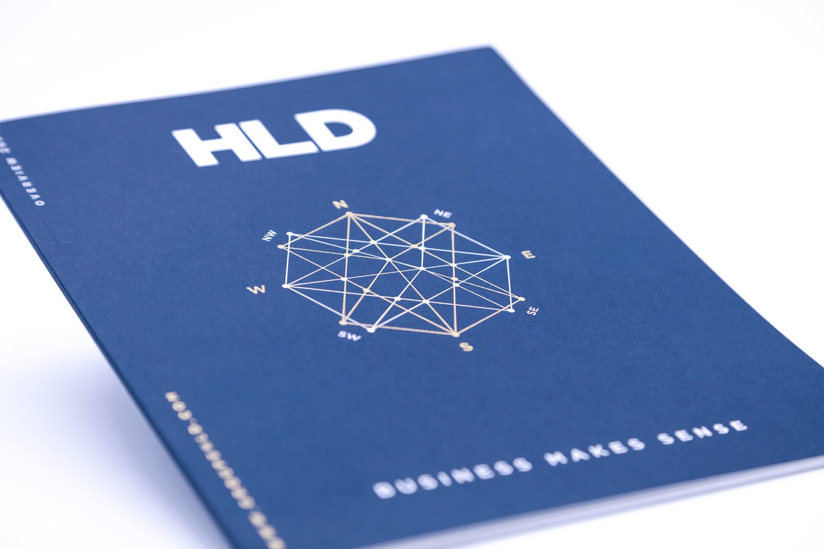 Un livret bleu avec HLD en lettres blanches en gras en haut, un dessin géométrique de compas en or au centre, et les mots BUSINESS MAKES SENSE en bas.