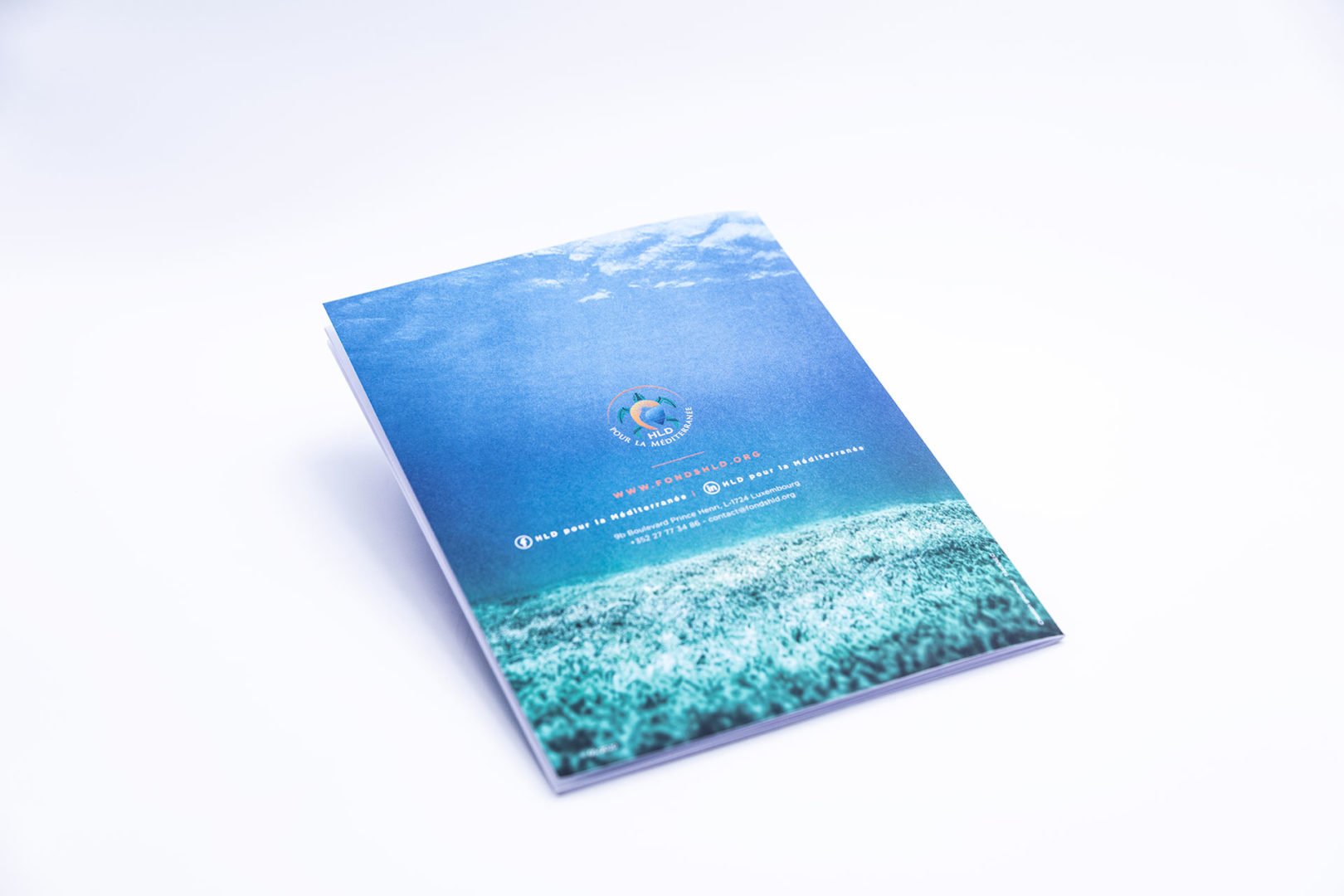Livre dont la couverture représente une scène sous-marine, avec de l'eau bleue et un fond sablonneux. Le centre est orné d'un logo circulaire et d'un texte blanc. Le livre est posé sur une surface blanche.