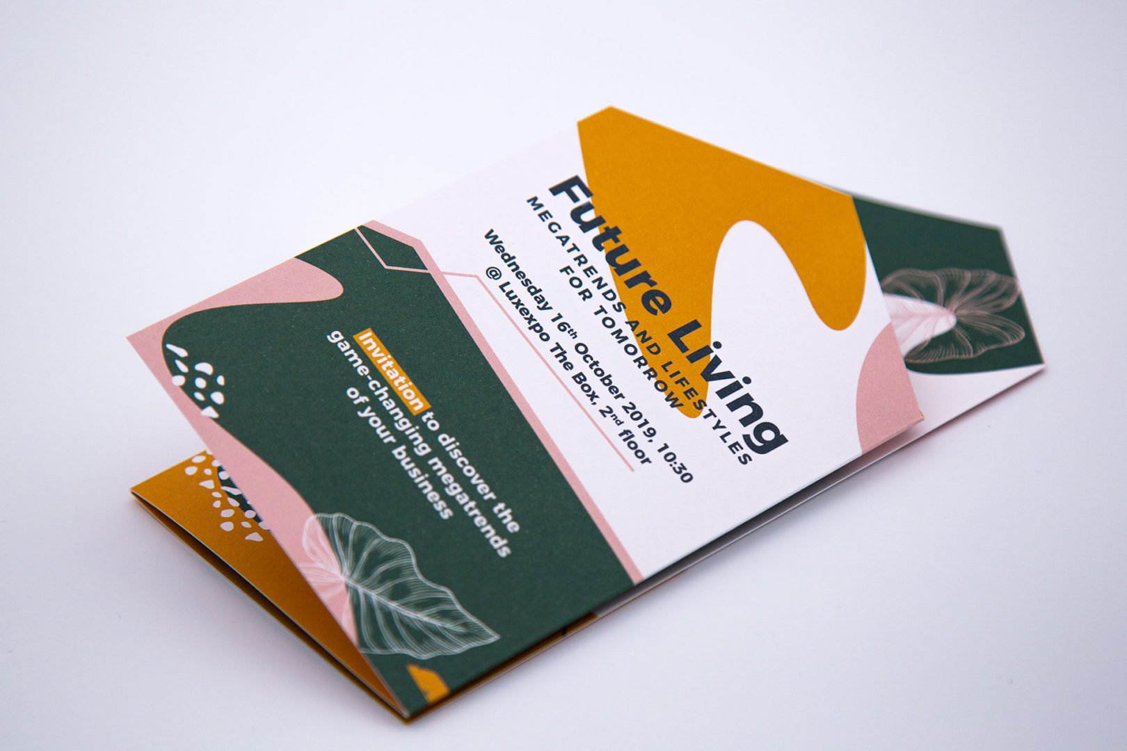 Carte d'invitation à un événement pliée avec des formes abstraites modernes en vert, jaune moutarde et rose vif. La carte porte la mention Future Living MEGA TRENDS, LIFESTYLES et fournit des détails sur l'événement ainsi qu'une invitation à découvrir de nouvelles opportunités commerciales.