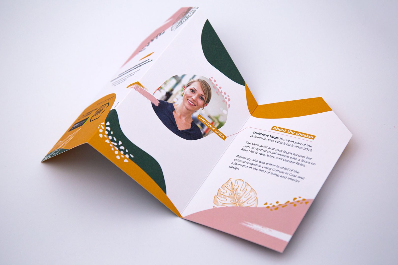 Brochure pliée en couleurs avec des accents verts, jaunes, roses et blancs, présentant une photo circulaire d'une femme et un texte sur un conférencier sur le panneau intérieur, ouverte sur une surface blanche.