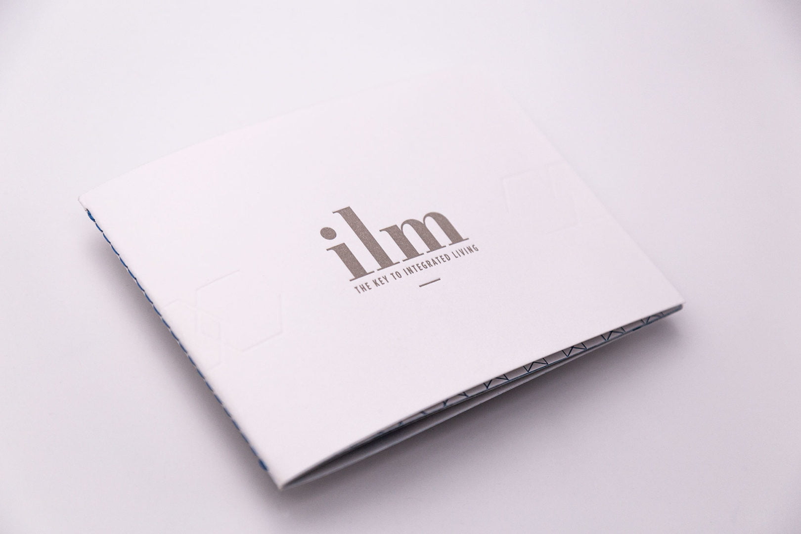 Sur un fond blanc uni, une brochure blanche de forme carrée avec le ilm et le slogan THE KEY TO INTEGRATED LIVING imprimé en gris argent sur la couverture.