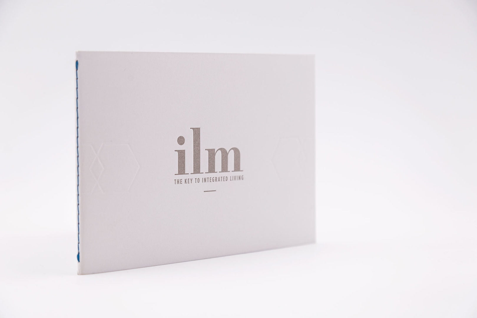 Un livret blanc se tenant debout avec le texte argenté ilm et le sous-titre The Key to Integrated Living sur la couverture, sur un fond blanc uni.