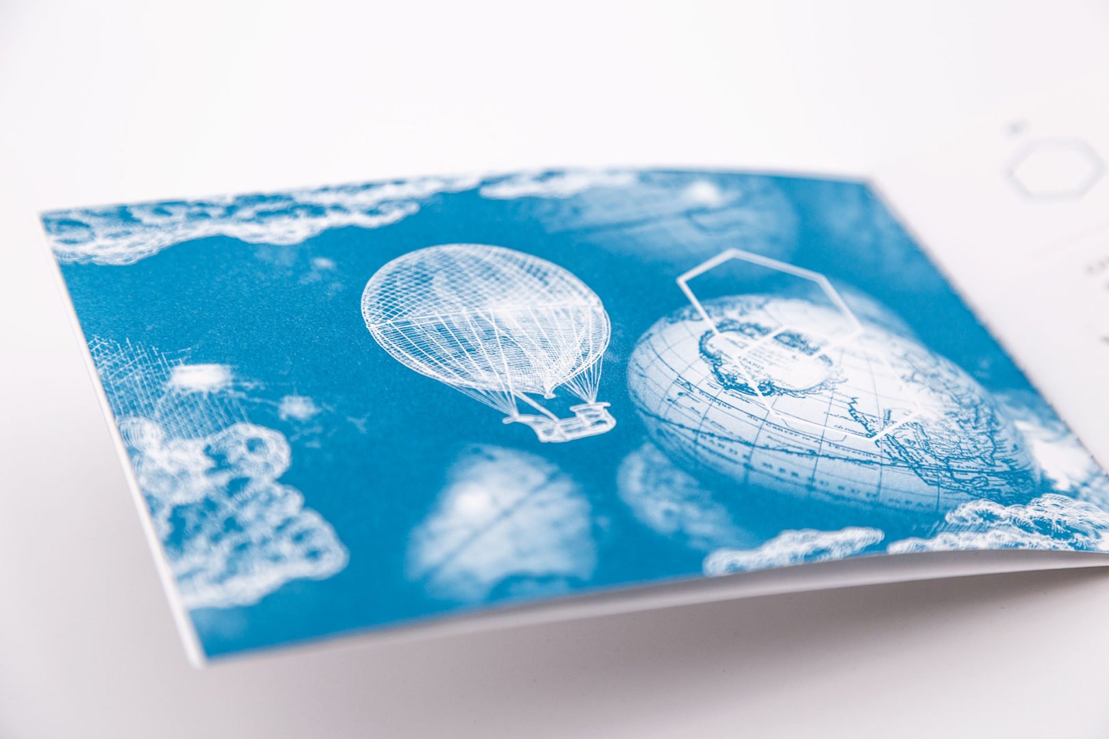 Gros plan sur une carte bleue et blanche illustrée d'une montgolfière, de nuages et d'un globe terrestre. Les illustrations ont un style vintage, proche du croquis. La carte repose sur une surface blanche.