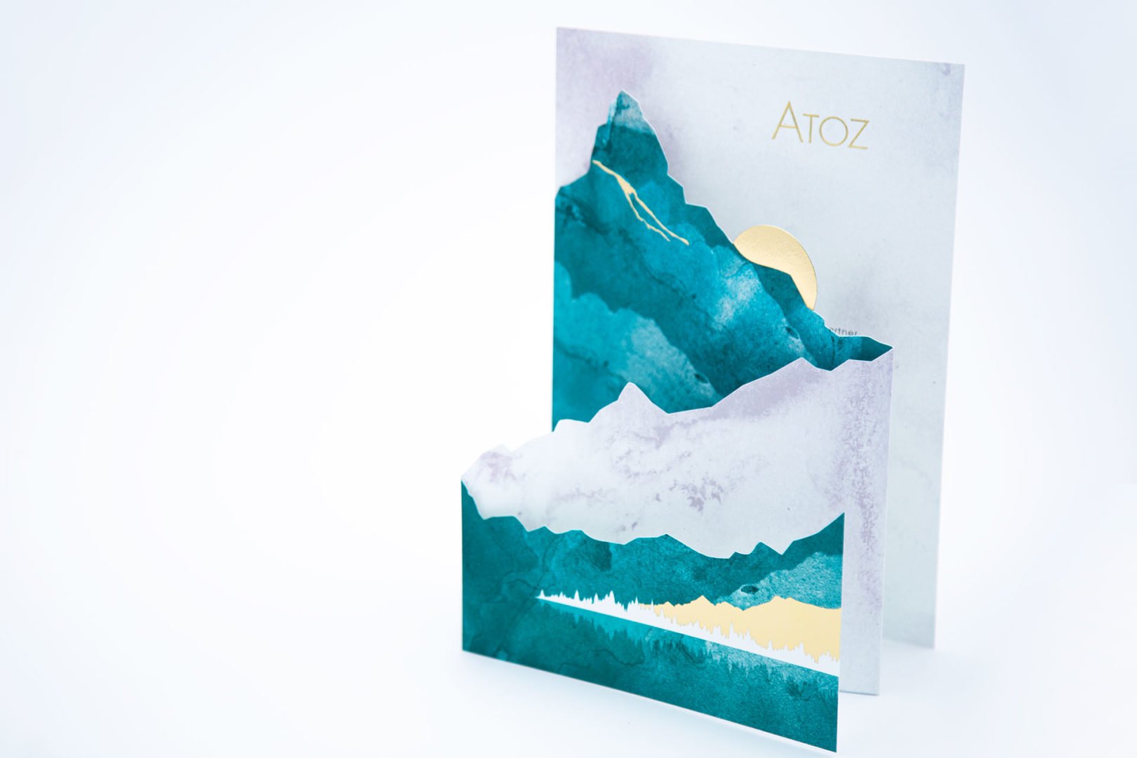 Carte décorative représentant des découpages superposés de montagnes bleues et blanches avec un soleil doré, debout sur un fond blanc. La carte porte l'inscription ATOZ en or en haut à droite.