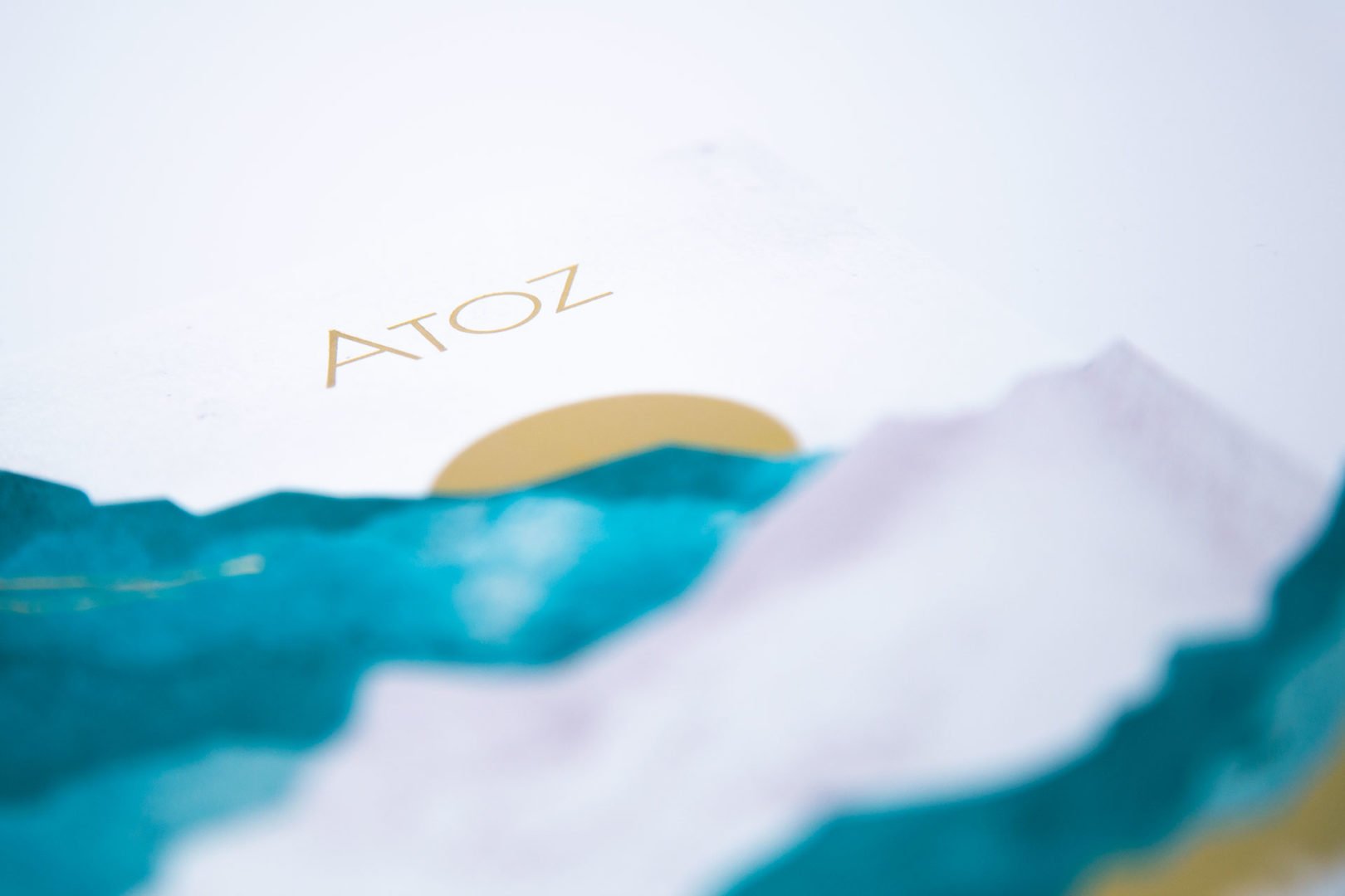 Gros plan d'un papier blanc portant le mot ATOZ en lettres dorées en haut, avec un dessin abstrait de montagnes bleues et un cercle doré en bas. La mise au point de l'image est douce et floue.