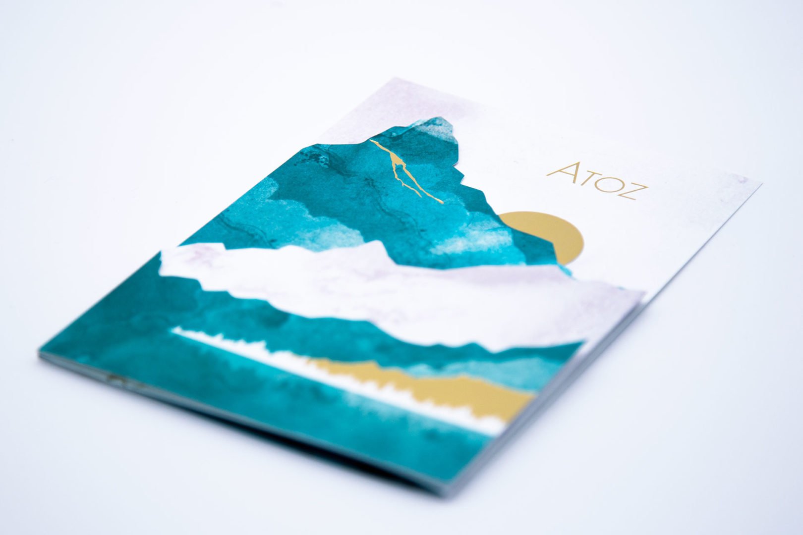 Un livret avec un dessin abstrait de montagne et de soleil dans des tons sarcelle, blanc et or, intitulé Atoz, placé sur une surface blanche.