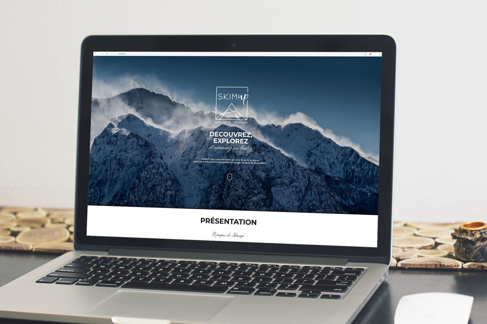 Un ordinateur portable posé sur une table en bois affiche un site web sur fond de montagne enneigée avec le texte Découvrez. Explorez. en français, ainsi qu'un logo et le mot Présentation visible en dessous.