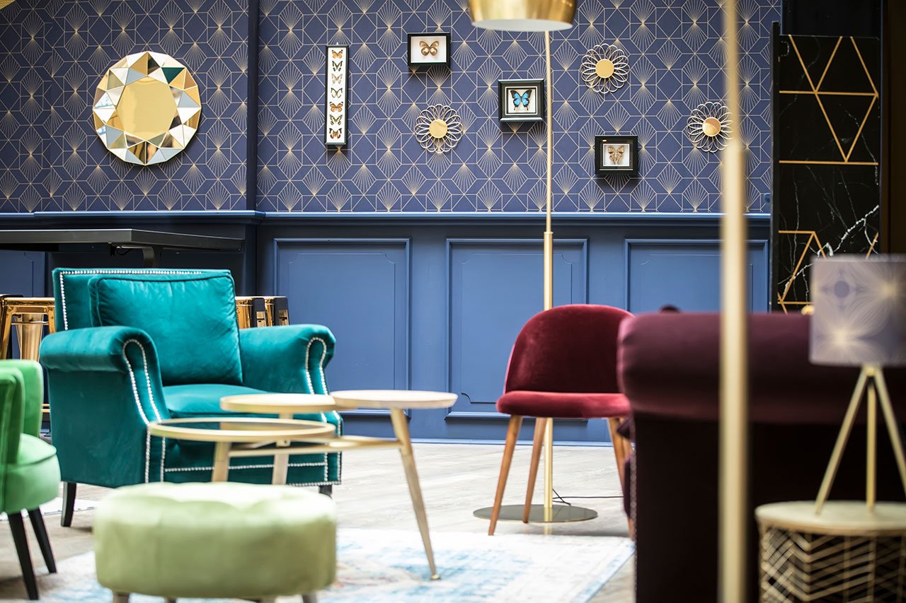 Un salon élégant avec des chaises colorées, un fauteuil sarcelle, une chaise marron, des tables rondes en bois, un ottoman vert et un mur bleu décoré de motifs géométriques, de miroirs et de papillons.