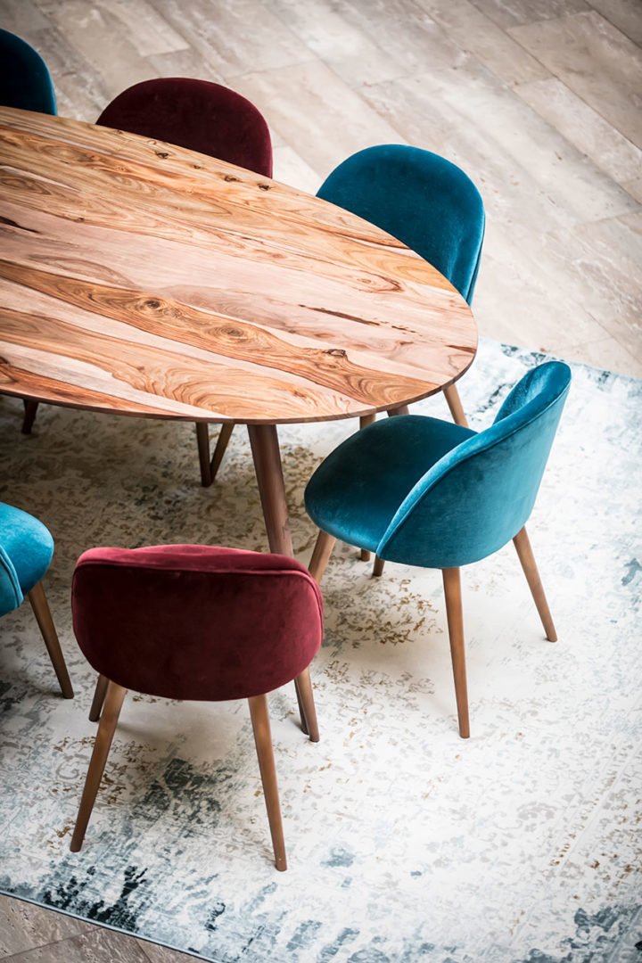 Une table ovale en bois entourée de chaises en velours, dont trois en sarcelle et une en bordeaux, est placée sur un tapis à motifs clairs sur un sol en bois.