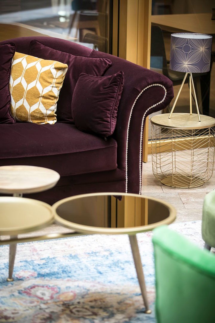Un coin de salon confortable avec un canapé en velours violet, un coussin géométrique jaune et blanc, des tables rondes dorées, une table d'appoint en fil de fer doré et une lampe moderne avec un abat-jour à motifs. Un tapis pastel et une chaise verte sont partiellement visibles.