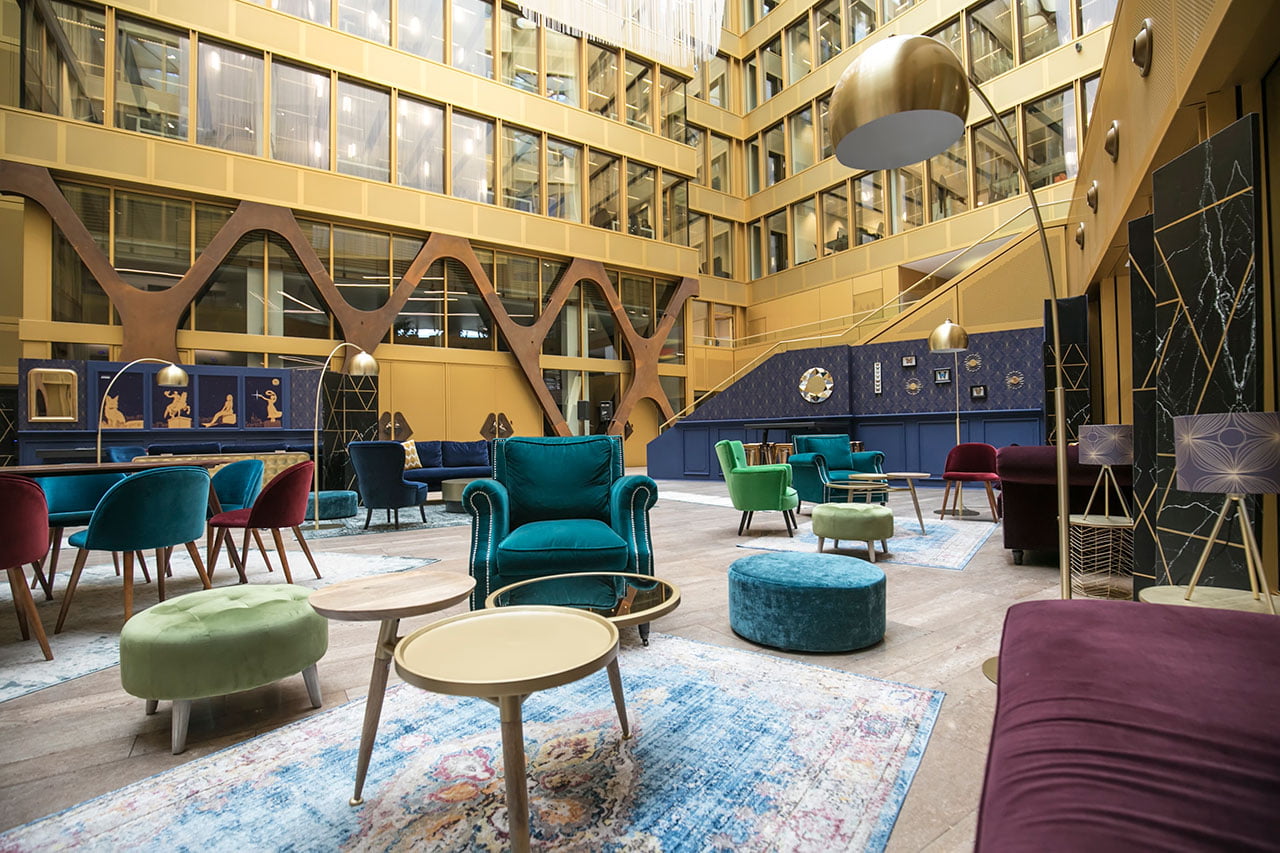 Un hall d'hôtel moderne avec des chaises en velours coloré, des tables rondes et des tapis à motifs. De grandes fenêtres et des accents dorés entourent l'espace, avec des décorations et des œuvres d'art bleues et dorées exposées sur les murs.