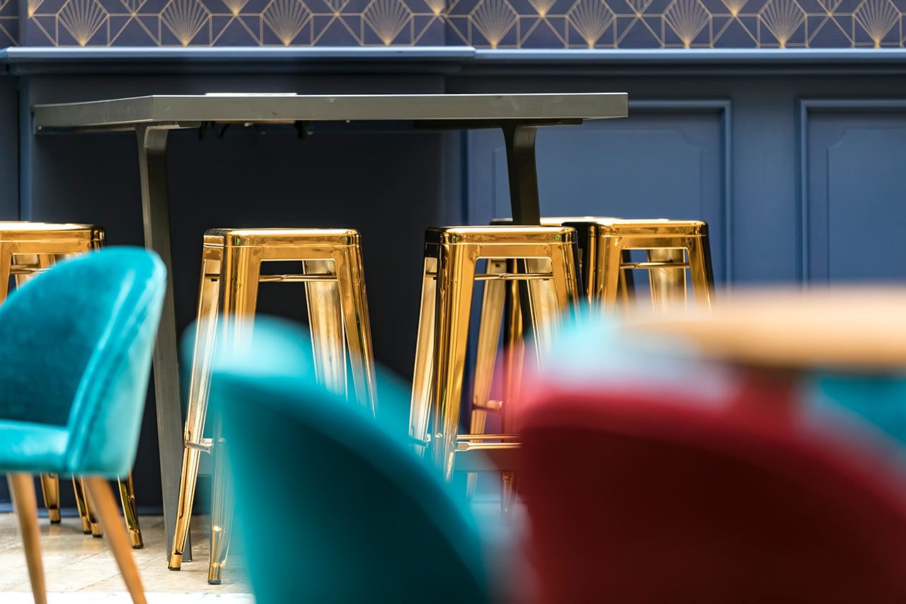 Un bar moderne avec des tabourets en métal doré brillant autour d'une table haute noire. Les chaises rembourrées sarcelles et rouges sont floues au premier plan, et le mur présente des motifs géométriques.