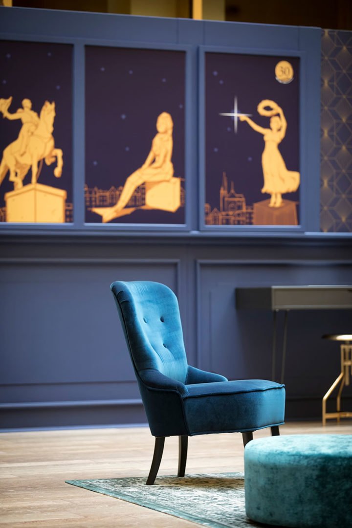 Une chaise bleue rembourrée est assise devant un mur décoré d'illustrations dorées de statues et de paysages urbains sur un fond bleu marine. Un petit ottoman rond et une partie d'un bureau sont également visibles.