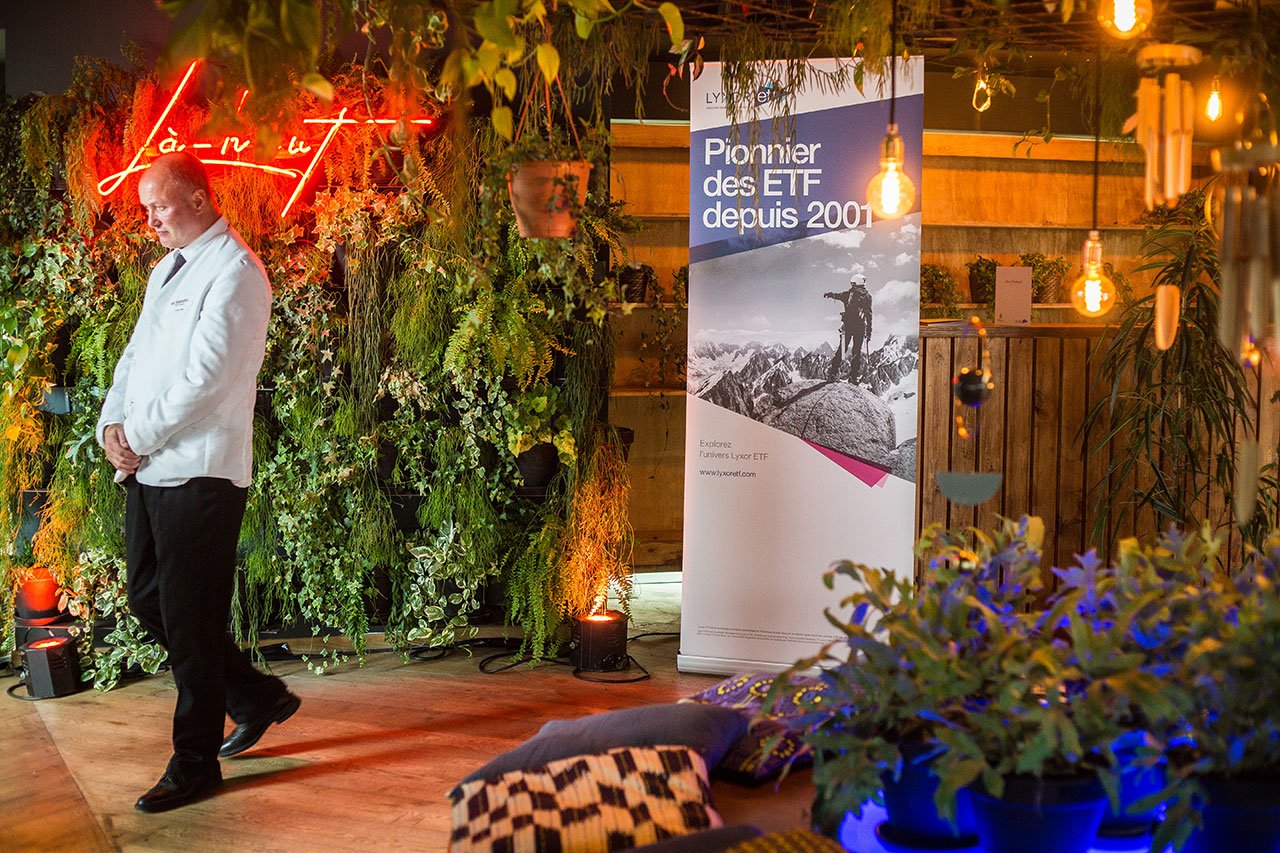 Un homme vêtu d'une veste blanche se tient près d'une verdure luxuriante et d'une enseigne au néon indiquant 
