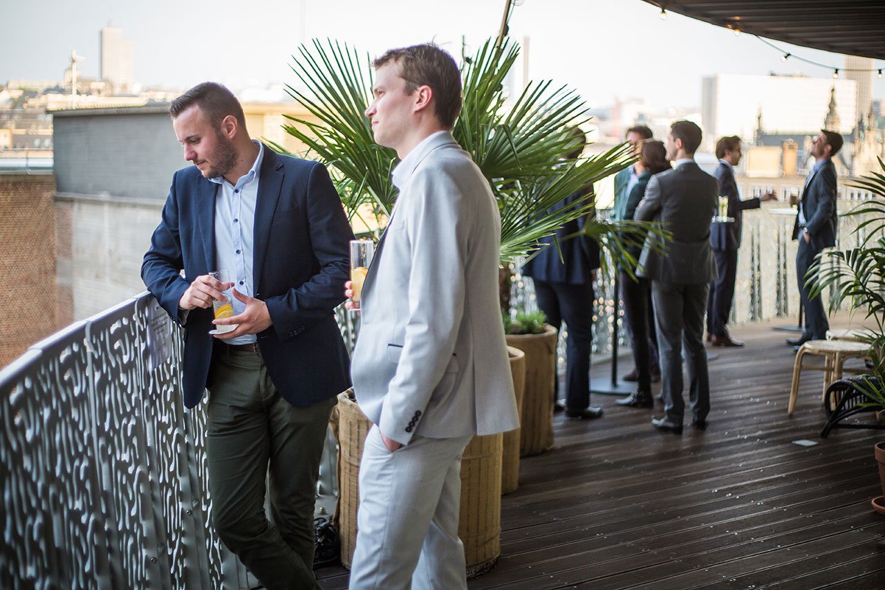 Des hommes en costume se tiennent debout et discutent sur un toit-terrasse avec des plantes en pot et des bâtiments urbains en arrière-plan. Deux hommes au premier plan discutent près d'une balustrade décorative.
