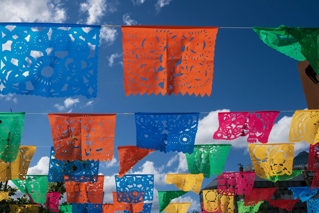 Des bannières colorées en papel picado sont suspendues à des ficelles dans un ciel bleu vif parsemé de nuages, créant ainsi une atmosphère de fête. Les bannières sont découpées de manière complexe dans différents motifs et couleurs, notamment orange, bleu, vert, jaune et rose.