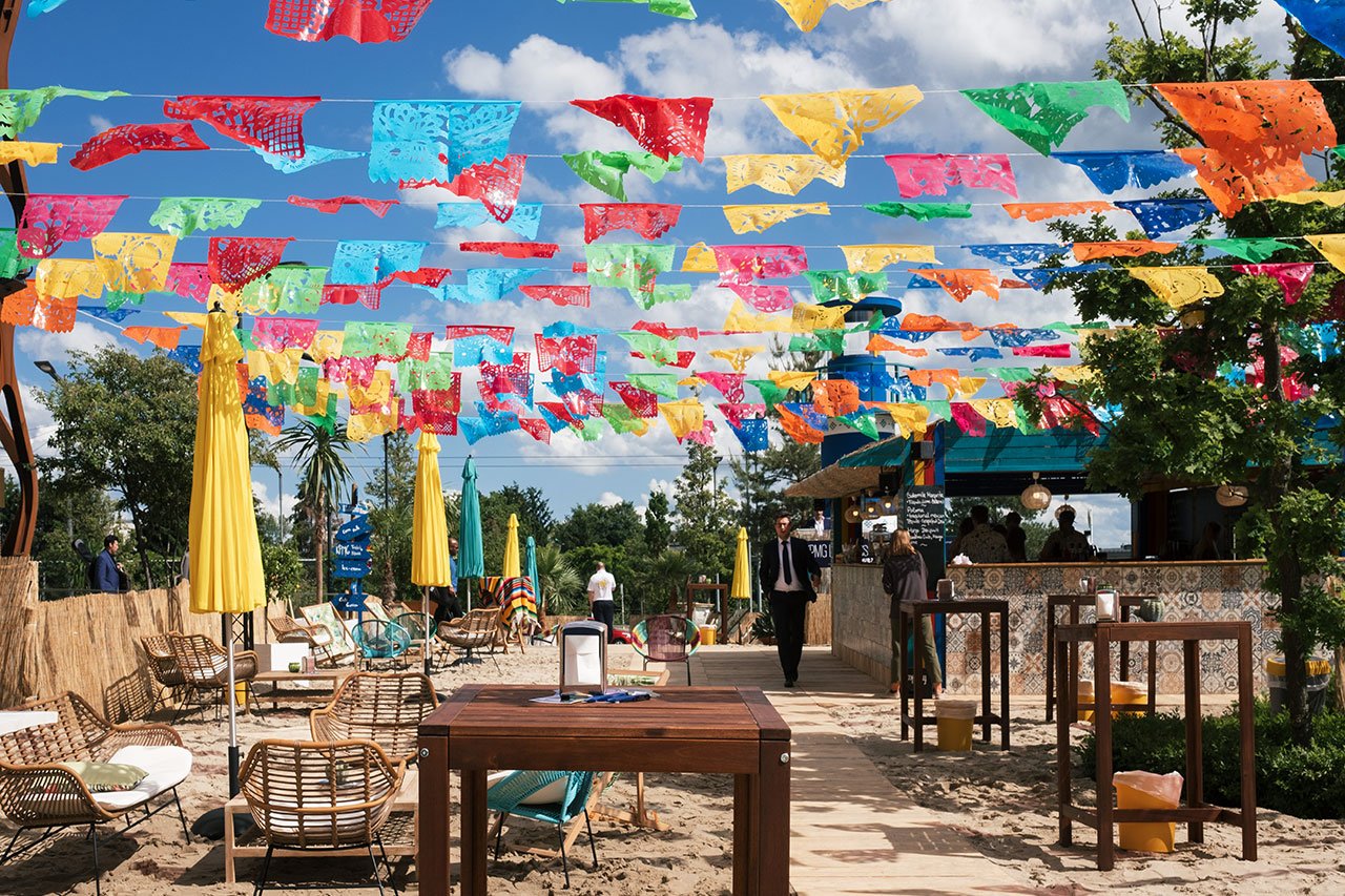 Des drapeaux en papier colorés sont suspendus au-dessus d'un bar de plage en plein air avec des parasols jaunes, des chaises en osier et des tables en bois sur le sable. Les gens sont rassemblés près du bar par une journée ensoleillée avec un ciel bleu et quelques nuages.