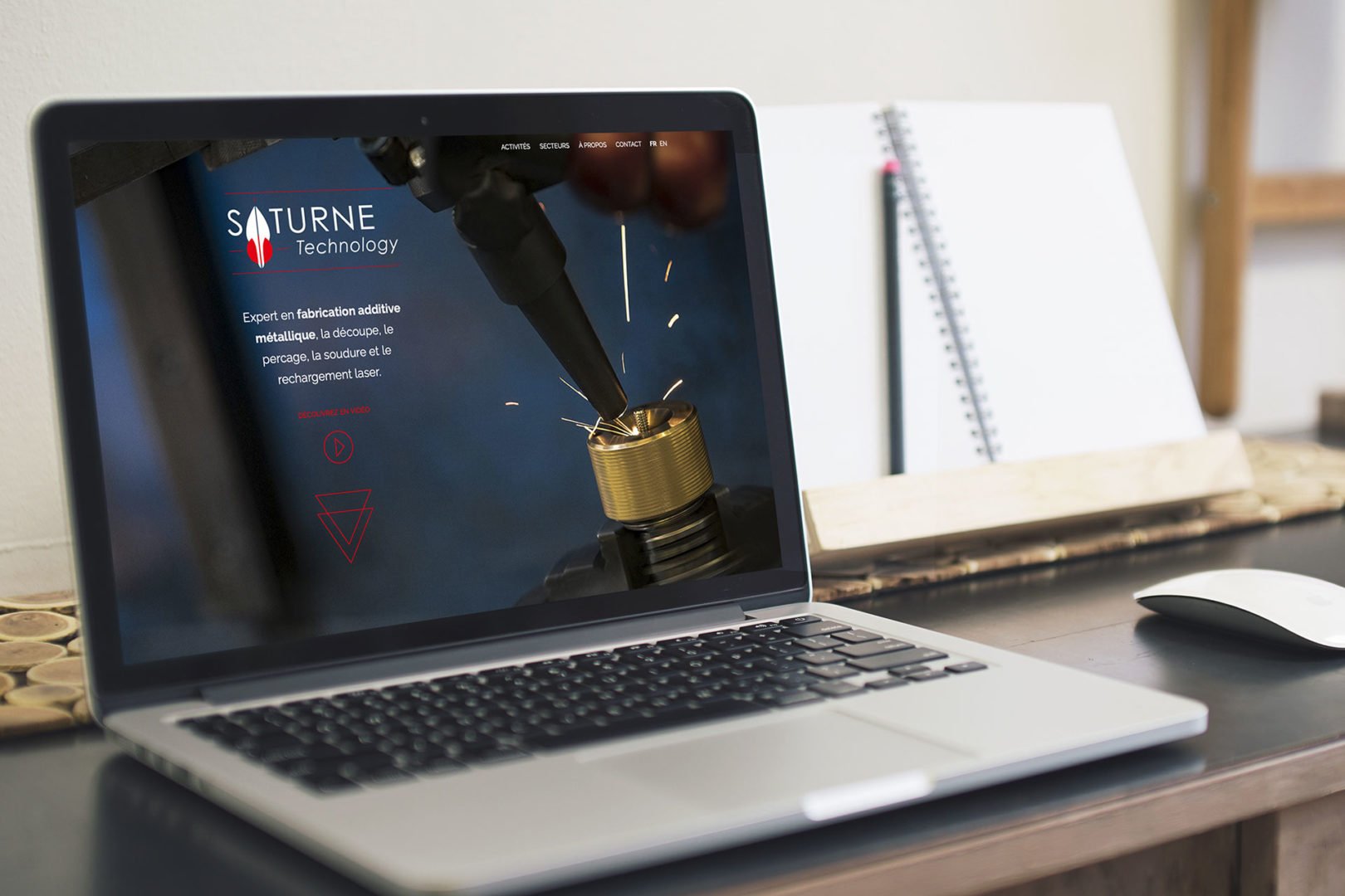 Un ordinateur portable posé sur un bureau affiche le site web de Saturne Technology, avec une image de fabrication additive métallique à l'aide d'un bras robotisé. Un ordinateur portable, un stylo et une souris sans fil se trouvent également sur le bureau.