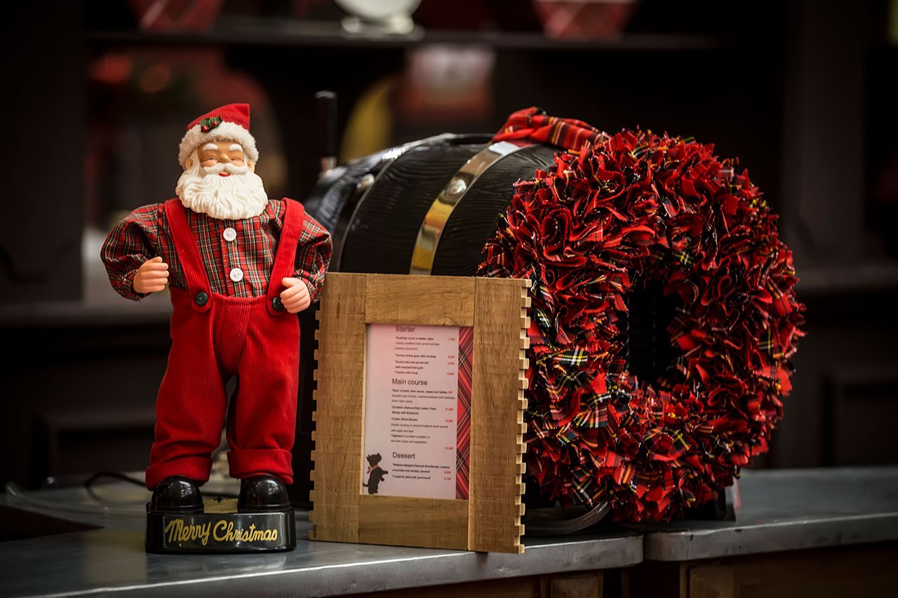 Une figurine du Père Noël en tissu écossais rouge se trouve à côté d'un menu encadré et d'une couronne de Noël rouge et noire, le tout disposé sur un plan de travail à l'arrière-plan flou.