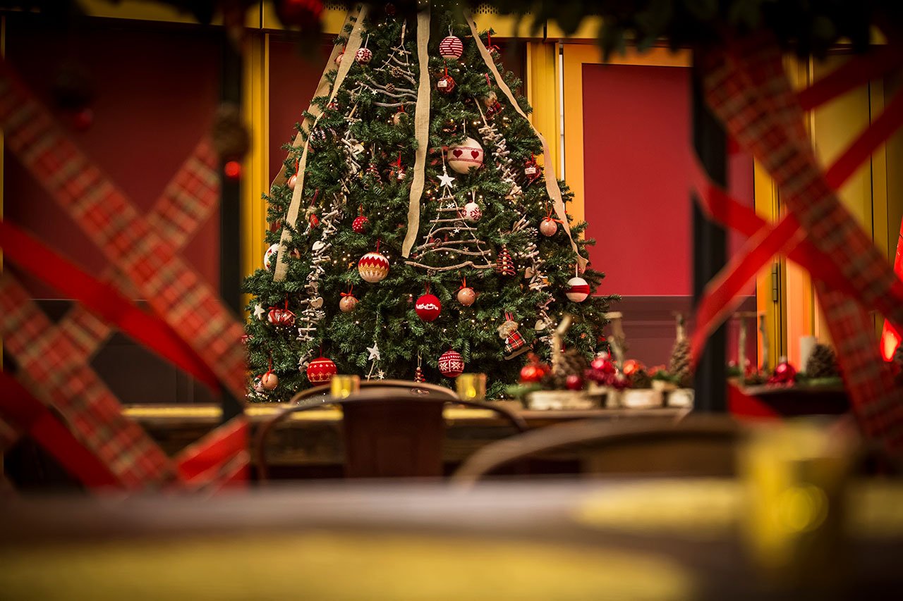 Un arbre de Noël décoré avec des ornements rouges et dorés se trouve à l'intérieur, avec des décorations rouges et dorées floues au premier plan et des tables placées devant l'arbre.