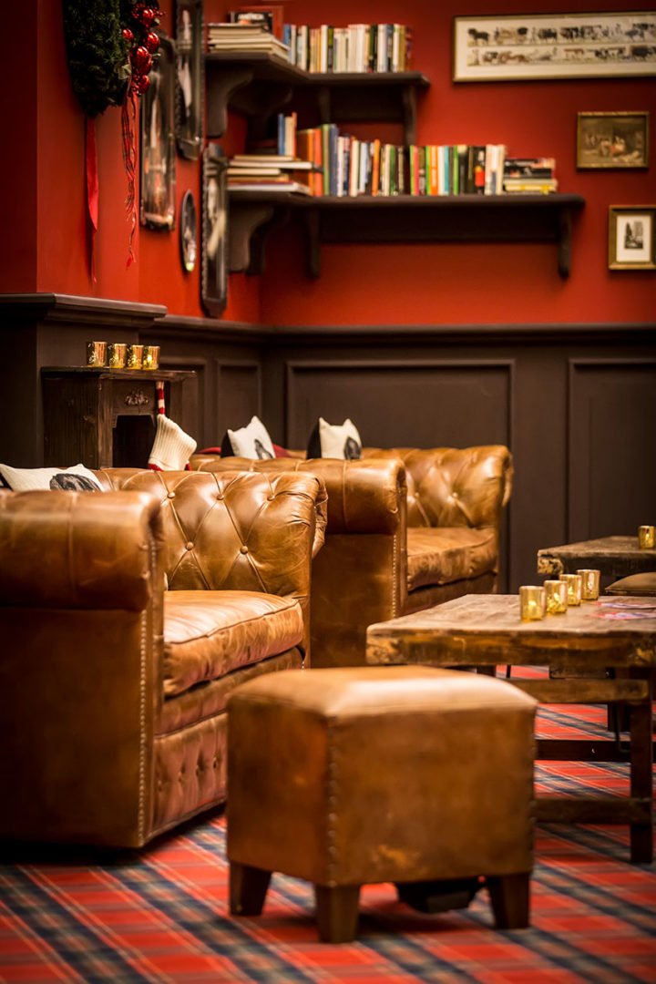 Un salon confortable avec des fauteuils et des ottomans en cuir brun, des tables en bois, une moquette à carreaux rouges et noirs, des murs rouges, des étagères avec des livres, des photos encadrées et des bougies, créant une atmosphère chaleureuse et accueillante.