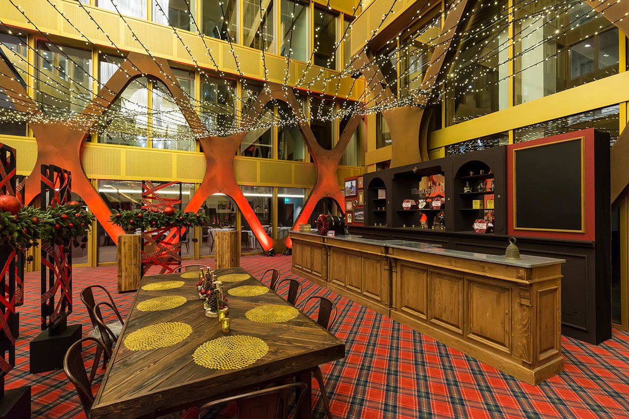 Un espace intérieur festif avec une longue table à manger en bois, des sets de table dorés, un bar en bois, un tapis à carreaux, des décorations de Noël et des guirlandes lumineuses au-dessus de la tête, le tout dans un bâtiment moderne aux accents jaunes et orange.