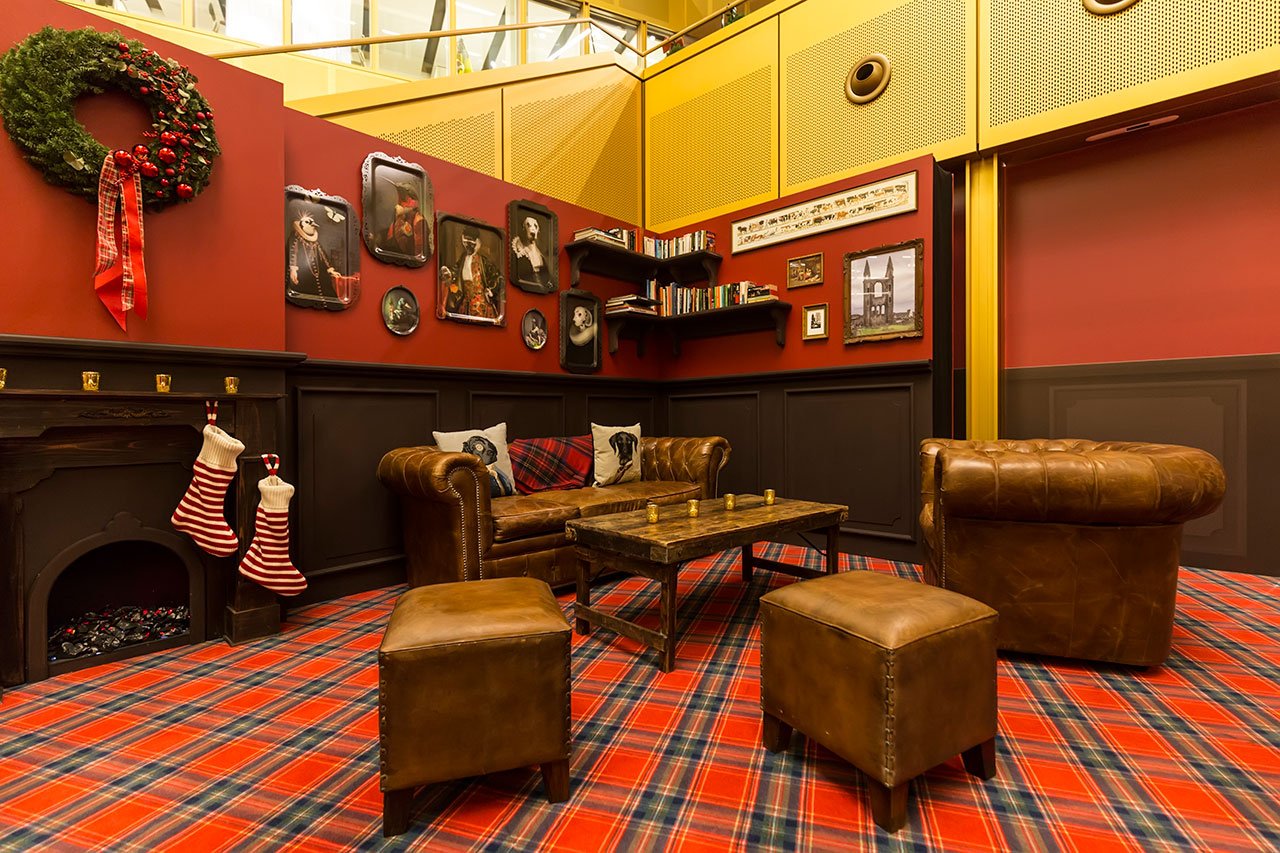 Un salon confortable avec un tapis à carreaux, un fauteuil et un canapé en cuir, une table basse, des bas suspendus à la cheminée, une couronne et des photos anciennes sur des murs rouges et bruns. Des étagères et des œuvres d'art encadrées décorent l'espace.