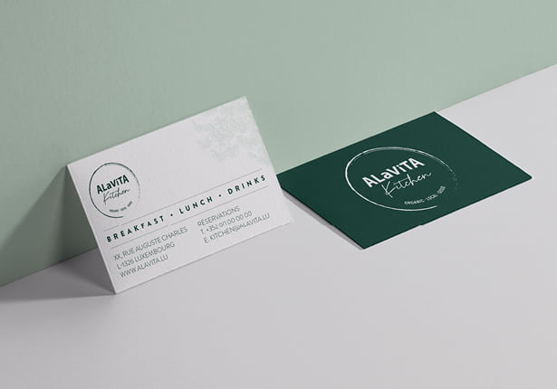 Deux cartes de restaurant pour la cuisine Alavita sont présentées sur une surface claire. L'une des cartes est blanche avec du texte, l'autre est vert foncé avec un logo circulaire blanc. Les deux cartes présentent un design épuré et moderne.