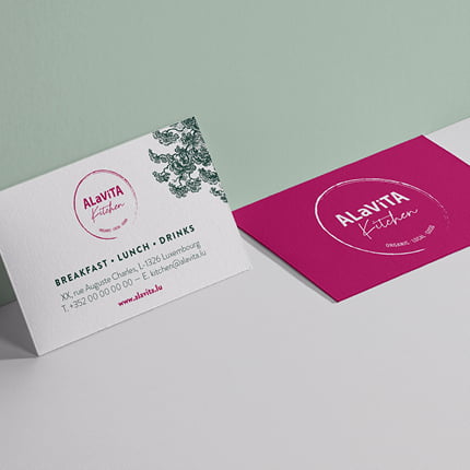 Deux cartes de visite pour ALaViTA Kitchen. L'une est blanche avec un motif floral vert et les coordonnées de la personne à contacter ; l'autre est magenta avec un logo blanc. Les deux cartes sont présentées sur une surface gris clair et verte.