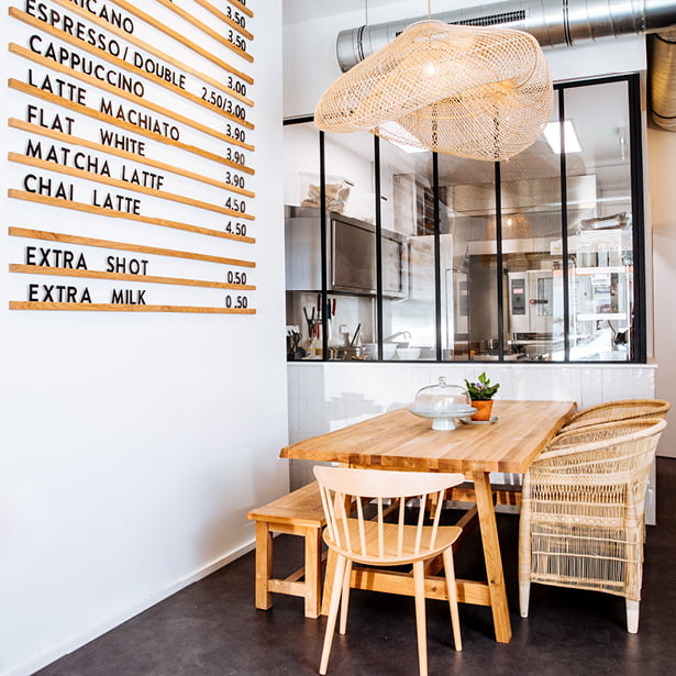 Un intérieur de café moderne et lumineux avec une table en bois et des chaises mixtes, une lampe suspendue tissée, une cuisine visible derrière une vitre et une carte murale indiquant les boissons et les prix du café.