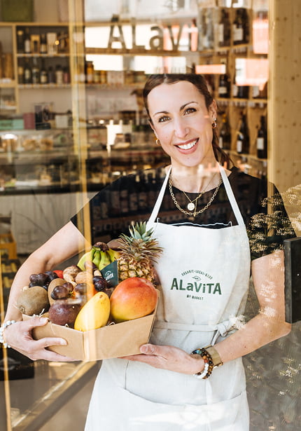 Une femme souriante portant un tablier blanc et des bijoux tient une boîte remplie de fruits frais assortis, debout à l'intérieur d'un magasin dont les étagères de produits sont visibles à l'arrière-plan.