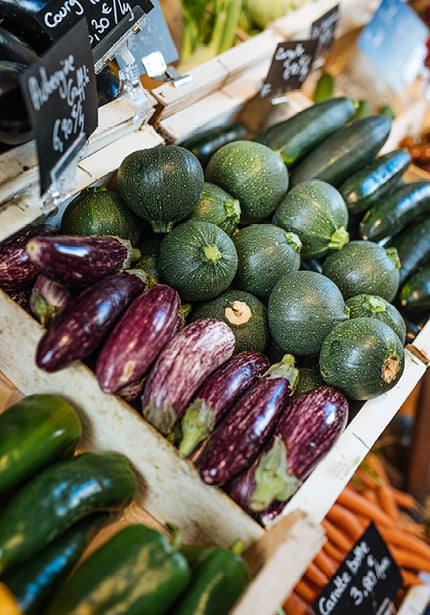 Un étalage de marché présentant des courgettes rondes, des aubergines violettes rayées et des courgettes vertes, toutes disposées dans des caisses en bois avec des panneaux de prix noirs.