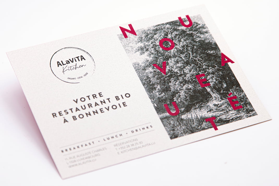 Carte postale portant le logo d'ALAVITA Kitchen, un texte en français faisant la promotion d'un restaurant biologique à Bonnevoie et une illustration détaillée d'un paysage en noir et blanc. Des lettres rouges en gras épellent NOUVEAUTÉ en diagonale sur le côté droit.