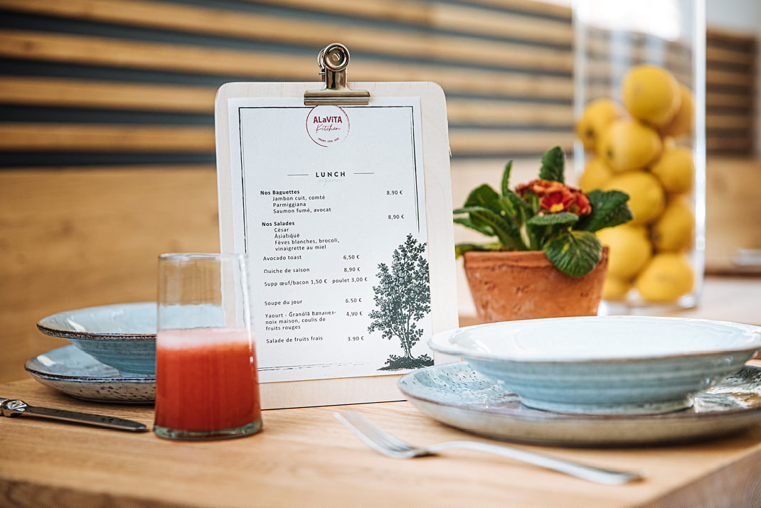 Un menu de déjeuner sur une planchette à pince est posé sur une table en bois à côté d'un verre de jus de fruit rouge, d'assiettes bleues empilées, d'une fourchette et d'un couteau, d'une fleur en pot et d'un vase en verre rempli de citrons.