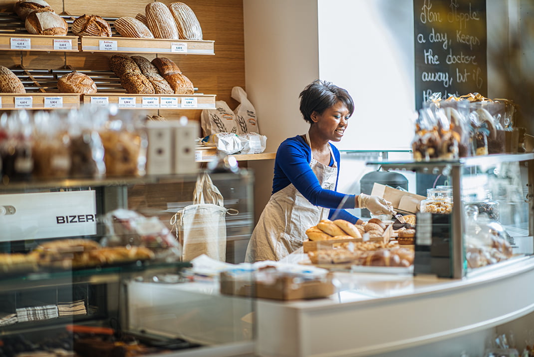 Une femme portant un tablier sourit alors qu'elle sert des pâtisseries derrière le comptoir d'une boulangerie, avec des étagères de pain et de produits emballés autour d'elle.