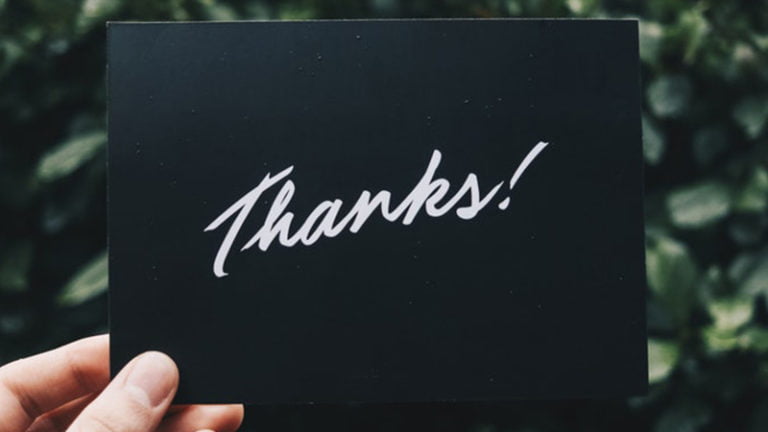 Une main tient une carte noire avec le mot "Thanks !" écrit en lettres cursives blanches. À l'arrière-plan, on aperçoit un feuillage vert flou.