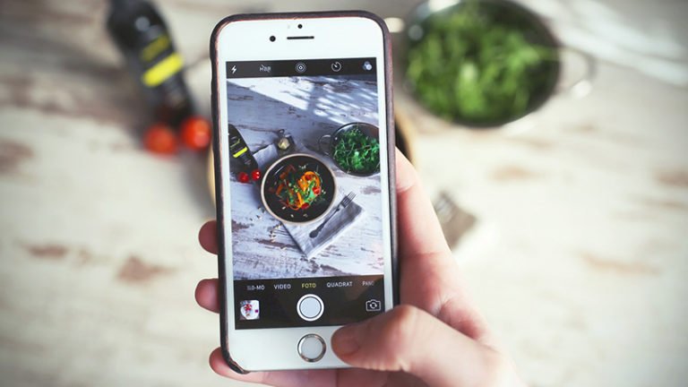 Une main tenant un smartphone prend une photo d'un bol de nourriture avec des légumes sur une table rustique, entouré d'ingrédients tels que des légumes verts, des tomates cerises et une bouteille en arrière-plan.