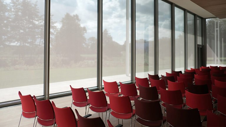 Des rangées de chaises rouges vides sont disposées dans une salle moderne dotée de grandes fenêtres allant du sol au plafond, permettant à la lumière naturelle d'entrer et offrant une vue sur les arbres et la verdure à l'extérieur.