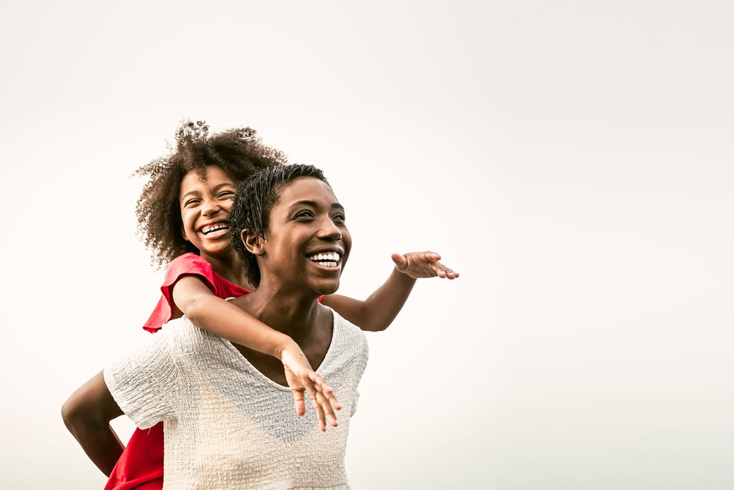 Une femme joyeuse fait faire un tour de cochon à une jeune fille souriante, toutes deux riant et s'amusant ensemble sur un fond clair et uni.