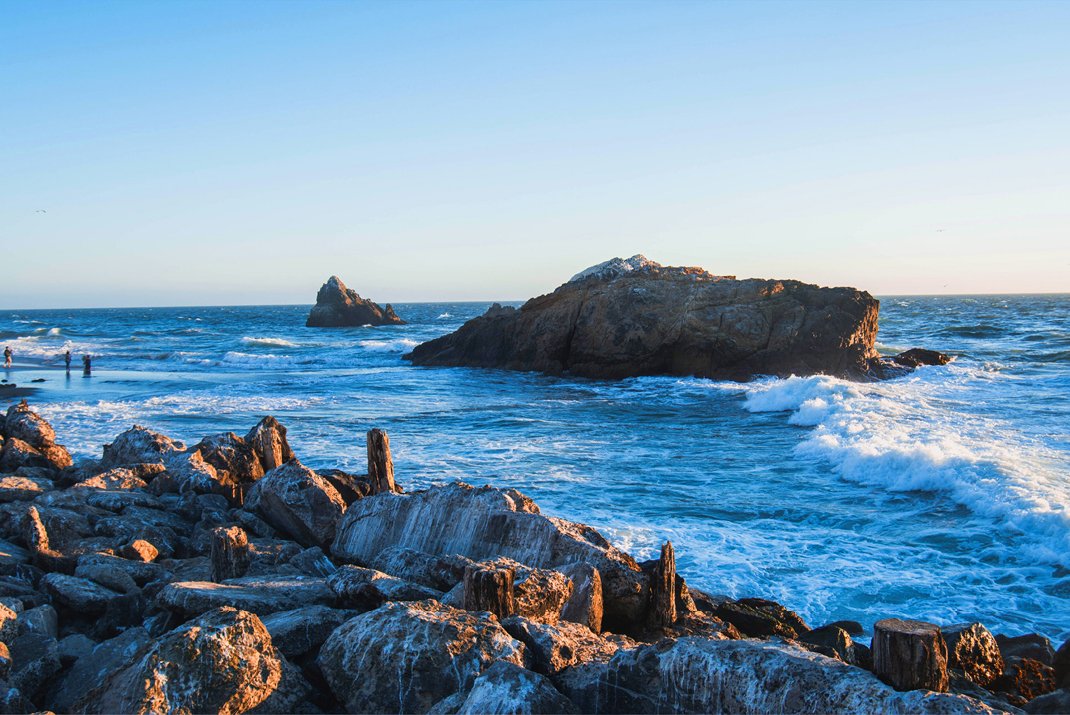 Côte rocheuse avec de gros rochers au premier plan, vagues s'écrasant contre les rochers dans l'océan, et ciel bleu clair au coucher ou au lever du soleil.