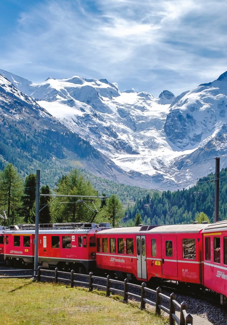 Un train de voyageurs rouge vif traverse un paysage alpin verdoyant au Luxembourg, passant devant de grands pins et des montagnes enneigées, sous un ciel bleu - une scène digne d'un article de magazine sur le style de vie.