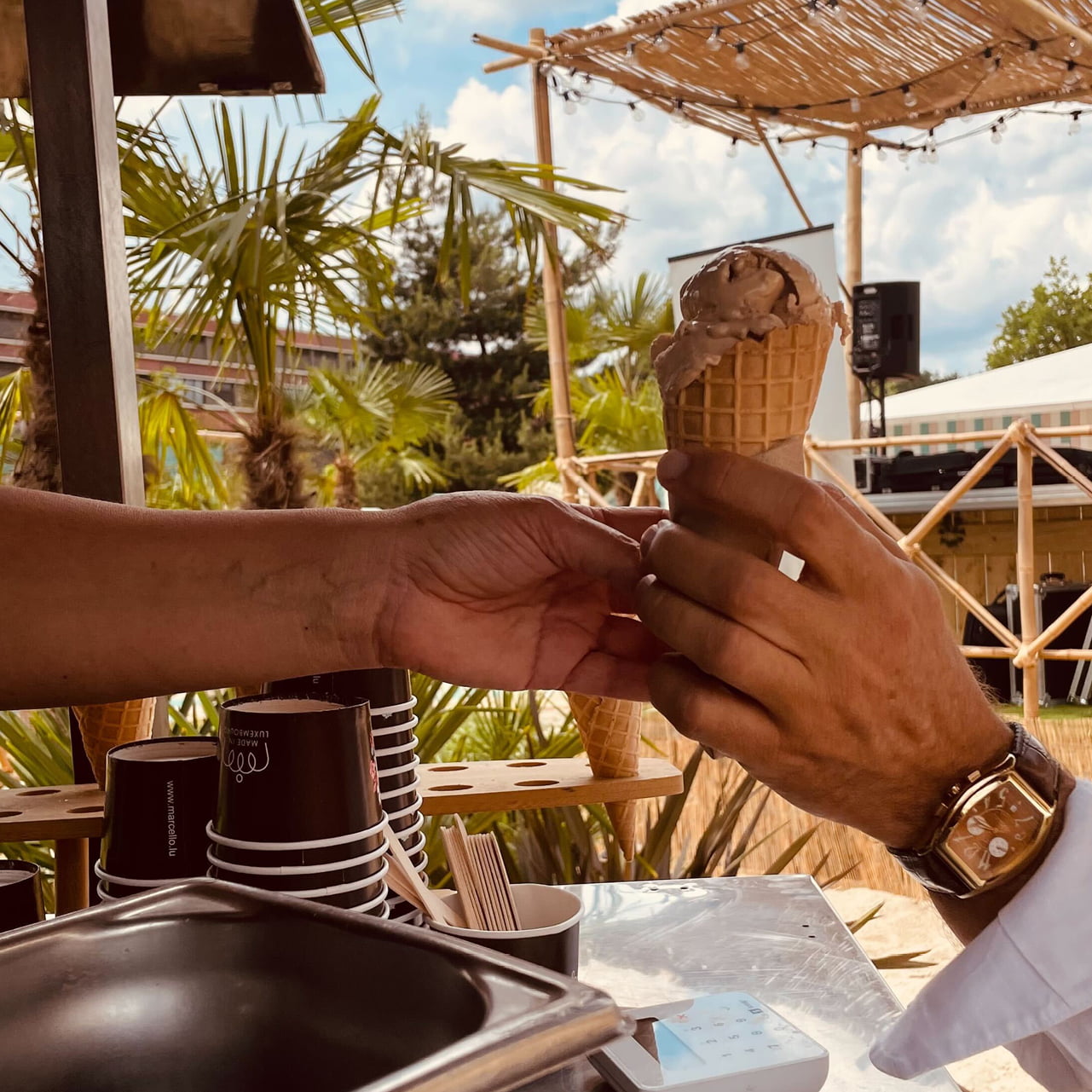 Une personne tend un cornet de glace à une autre personne portant une montre à un stand en plein air avec des palmiers, des coupes empilées et un ciel ensoleillé et partiellement nuageux en arrière-plan.