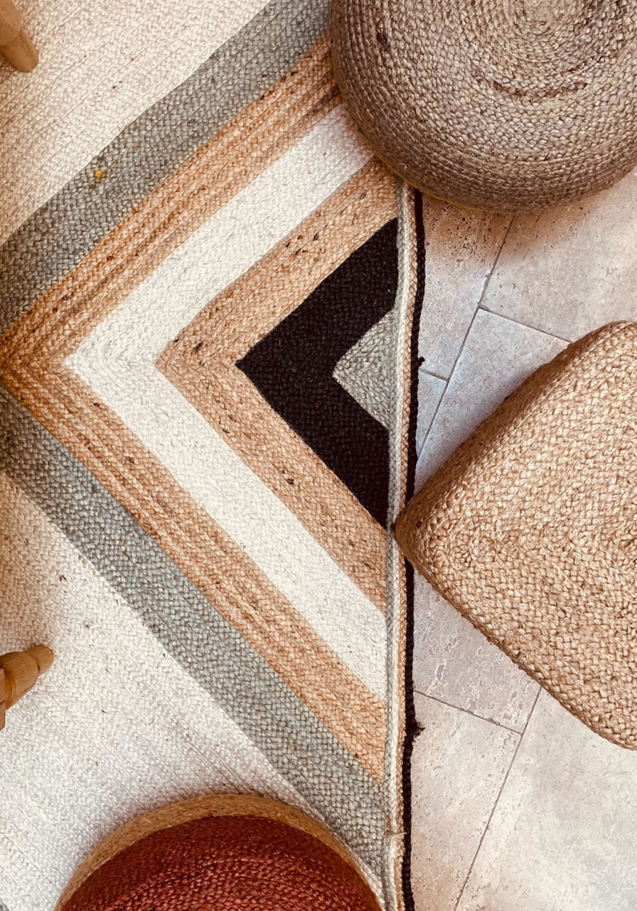 Un tapis géométrique à motifs chevron beige, beige, gris et noir est entouré de tabourets et de chaises tressés sur un sol carrelé. Les formes et les textures créent une atmosphère chaleureuse et terreuse.