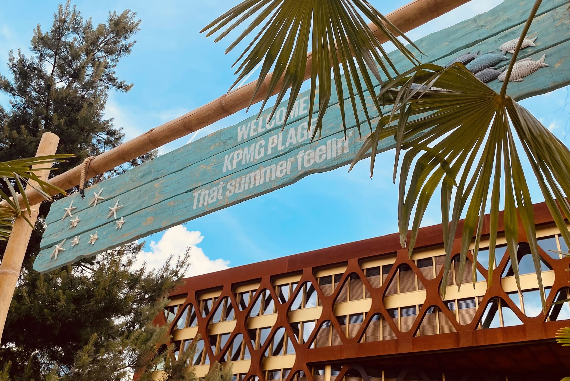 Un panneau en bois à thème tropical indique Welcome KPMG PLACE Thai summer feeling avec des feuilles de palmier au premier plan et un bâtiment moderne à l'arrière-plan sous un ciel bleu.