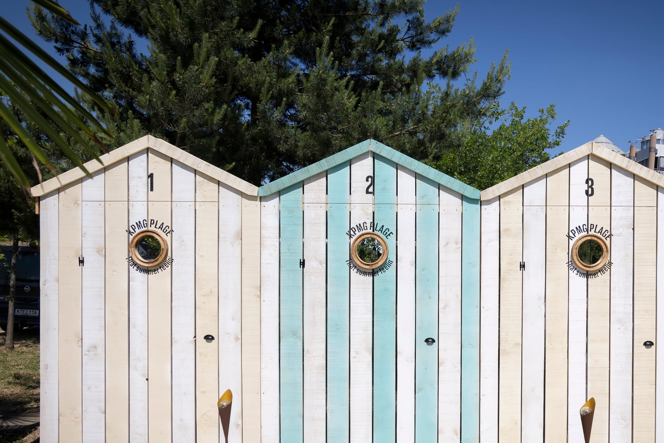 Trois cabines de plage en bois avec des toits en pointe, étiquetées 1, 2 et 3 ; la cabine du milieu est peinte en bleu tandis que les autres sont blanches. Chaque cabine est dotée d'une fenêtre ronde et des arbres sont visibles à l'arrière-plan.