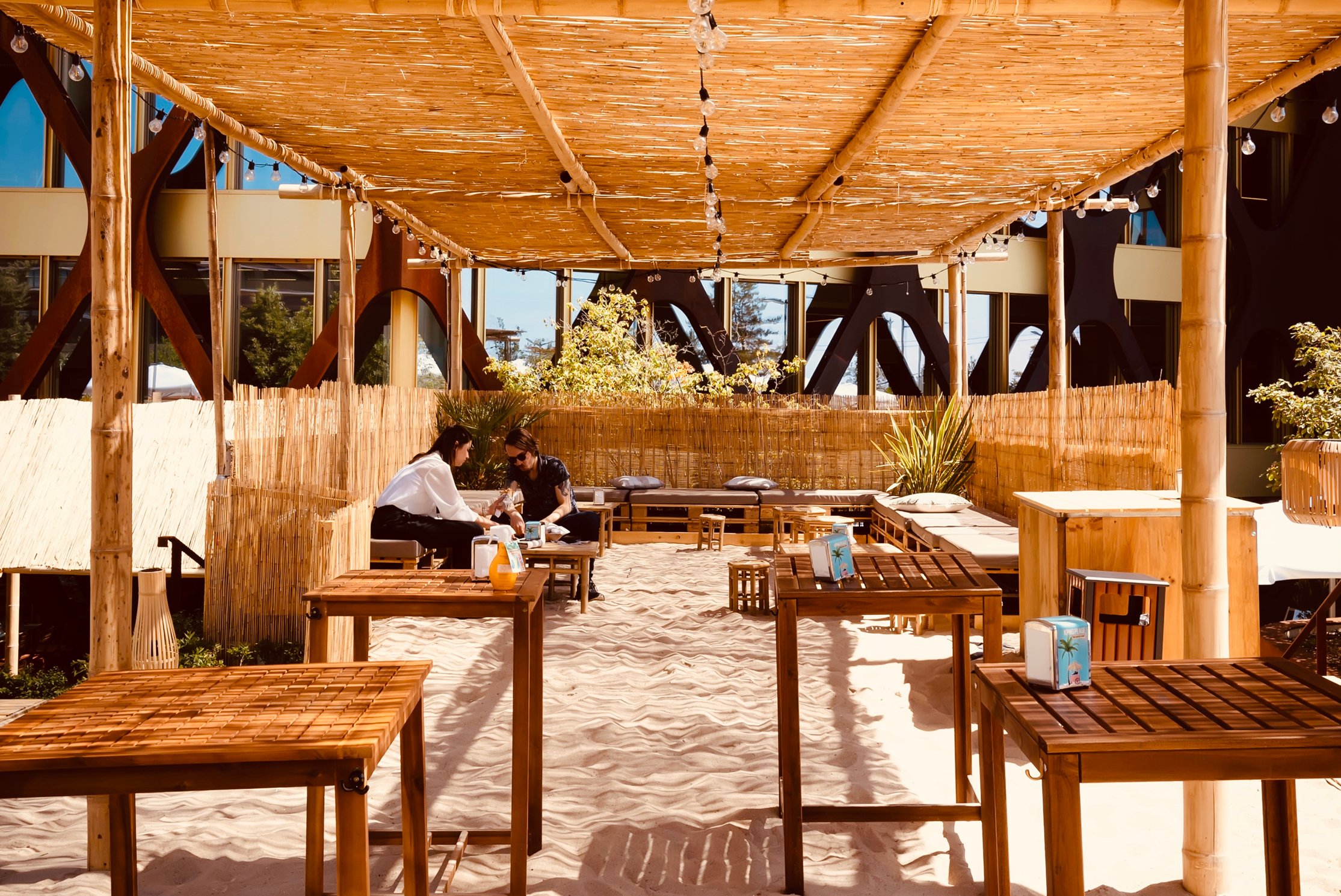 Café en plein air avec toit en bambou et sol en sable, tables et chaises en bois, deux personnes discutant à une table, entourées de plantes et de guirlandes lumineuses, créant une atmosphère décontractée, semblable à celle de la plage.