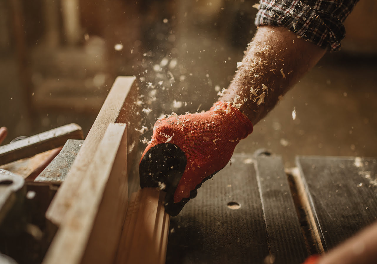 Une personne portant un gant rouge lisse une planche de bois à l'aide d'un rabot manuel, faisant voler des copeaux de bois dans un atelier.