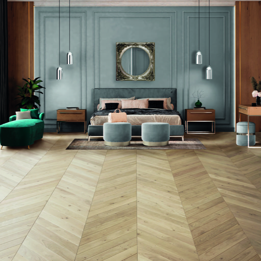 Chambre à coucher élégante avec un sol en bois clair à motifs de chevrons, des murs en lambris gris, un grand lit avec une literie neutre, deux tabourets gris, un fauteuil vert, des lampes suspendues et un miroir décoratif au-dessus du lit.