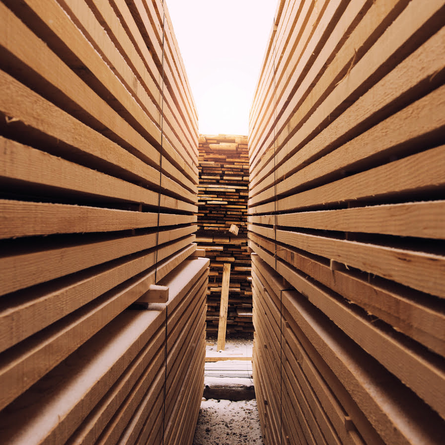 Des piles de planches de bois coupées sont disposées en rangées, formant un passage étroit entre elles, et la lumière du soleil brille au bout du couloir.