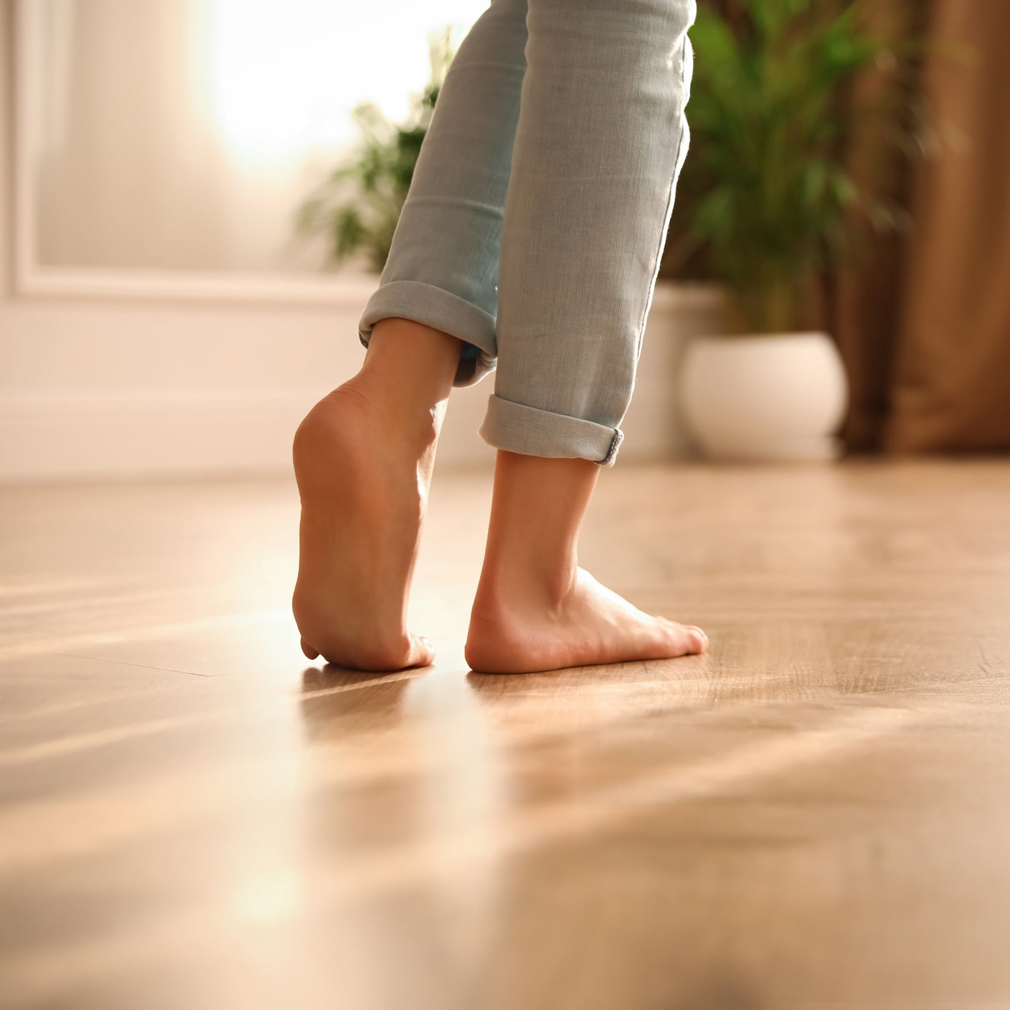 Une personne aux jeans retroussés se tient sur la pointe des pieds sur un plancher en bois à l'intérieur, avec des plantes vertes et une douce lumière naturelle en arrière-plan.